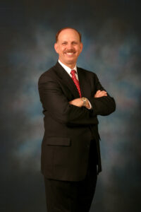 George Indest, of The Health Law Firm, standing for a headshot in a dark suit with red tie with arms crossed
